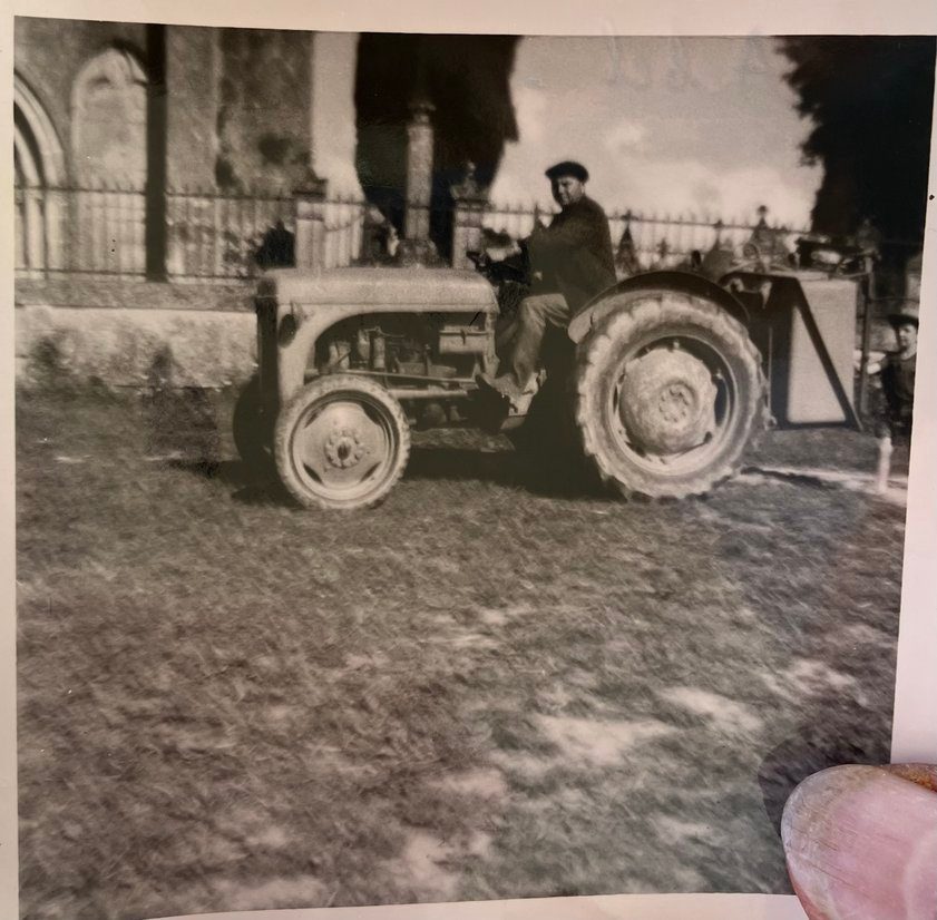 Photographie en noir et blanc d'un homme sur un tracteur, devant l'église et le cimetière